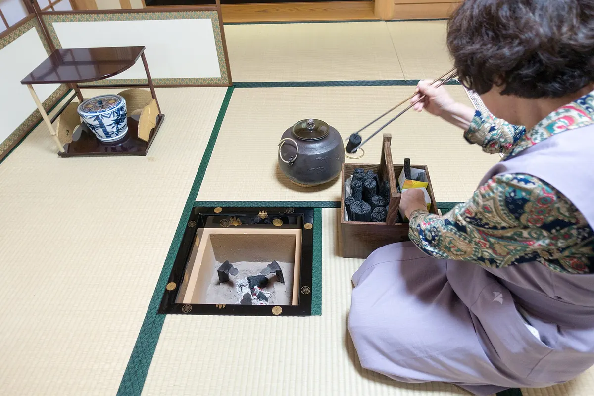 Žena v šeříkovém kimonu klečí na tatami vedle zapuštěného ohniště, dlouhými kleštěmi zvedá uhlíky a vedle ní stojí konvice a lakovaný tác s uhlím